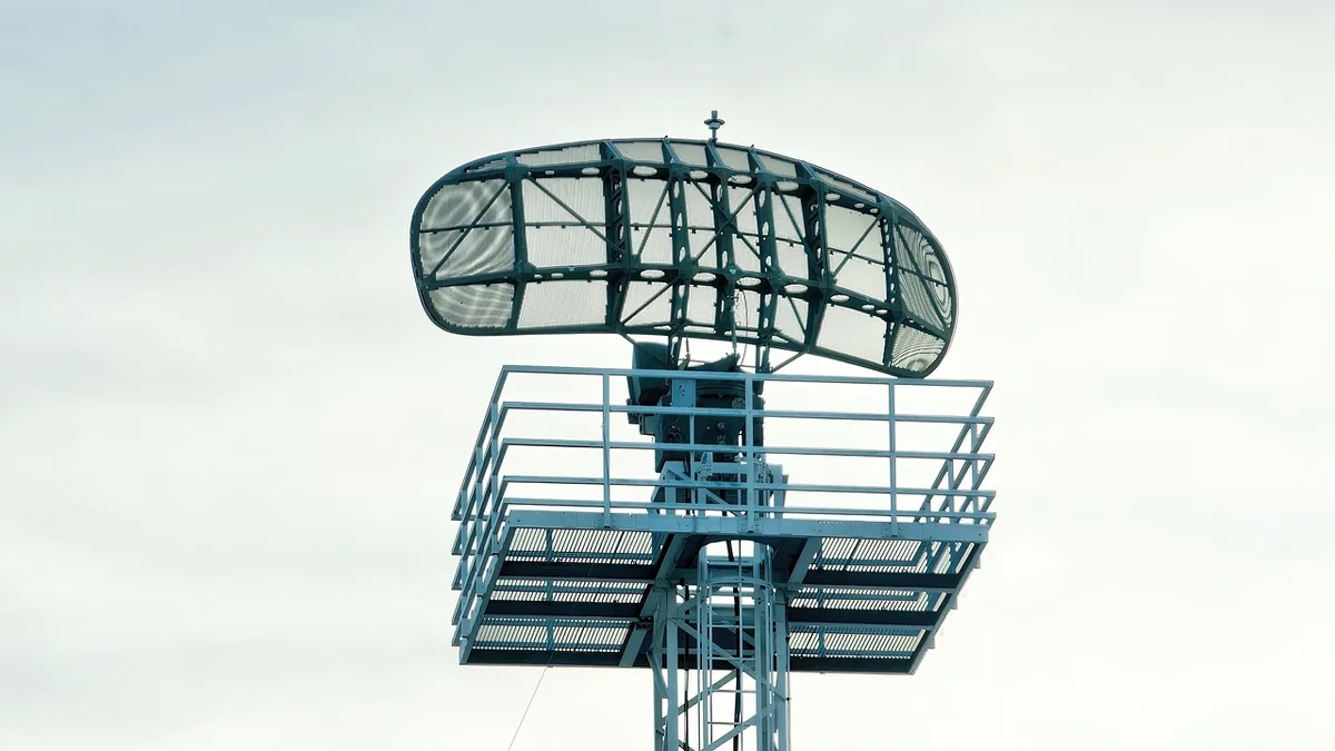 A large green radar antenna mounted on a metal tower against a cloudy sky.