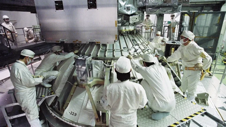 Technicians in white cleanroom suits assemble a large spacecraft component.
