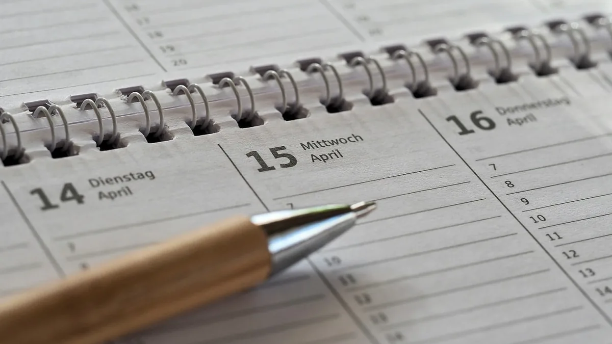A wooden pen rests on a spiral-bound calendar showing mid-April dates, symbolizing planning and timelines.