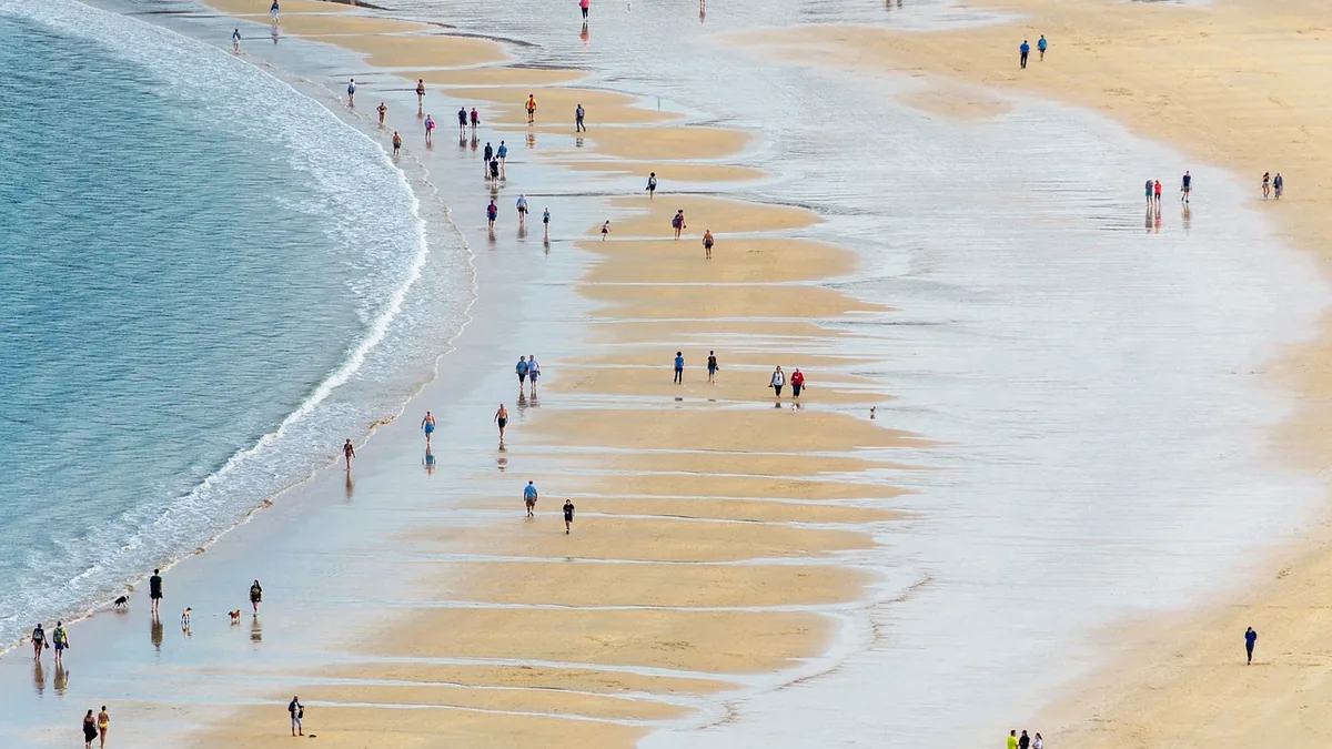 An overhead view of people walking on a sandy beach with geometric tidal ripples and wet sand reflections.