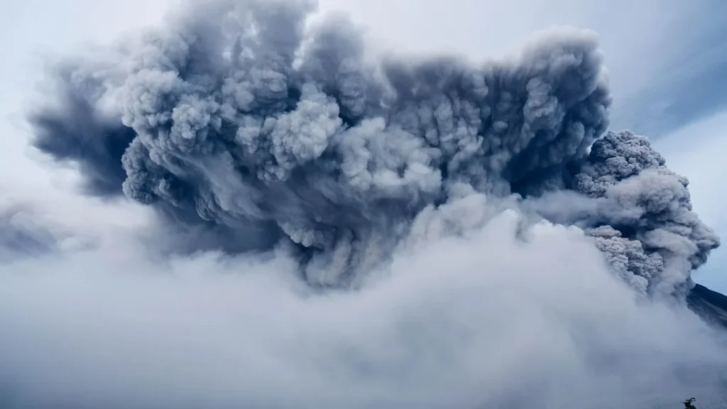 A massive cloud of dark grey volcanic ash billows into the sky above low clouds.