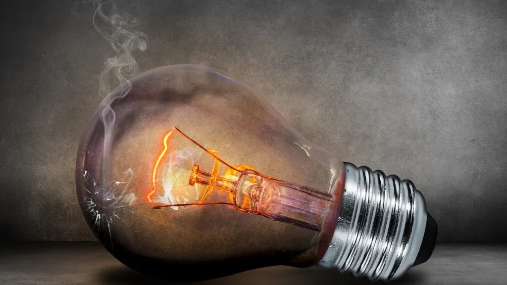 A cracked, smoking light bulb with a glowing filament lies on a dark surface.