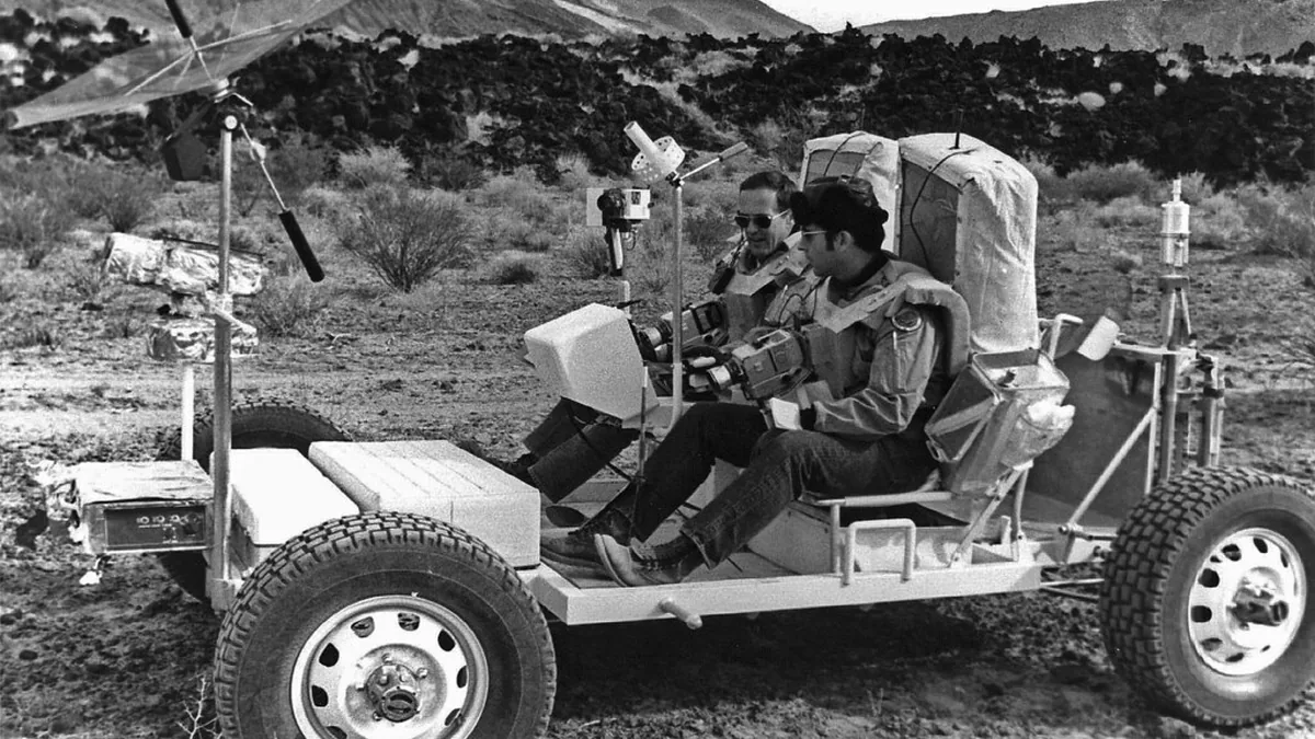 Astronauts in space suits sit in a lunar rover prototype during training in a desert landscape.