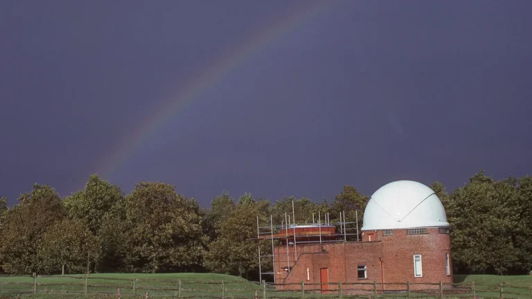 Red brick observatory with a white dome under a colorful rainbow against a dark sky.