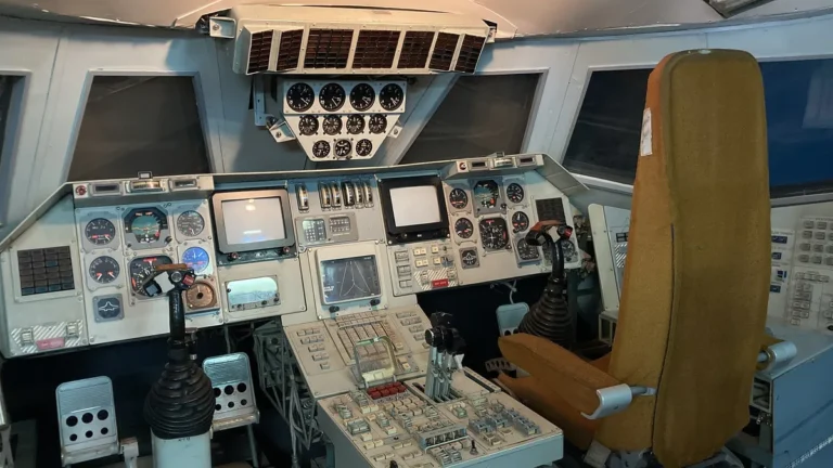 Vintage space cockpit with orange seat, grey control panels, joysticks, and CRT screens.