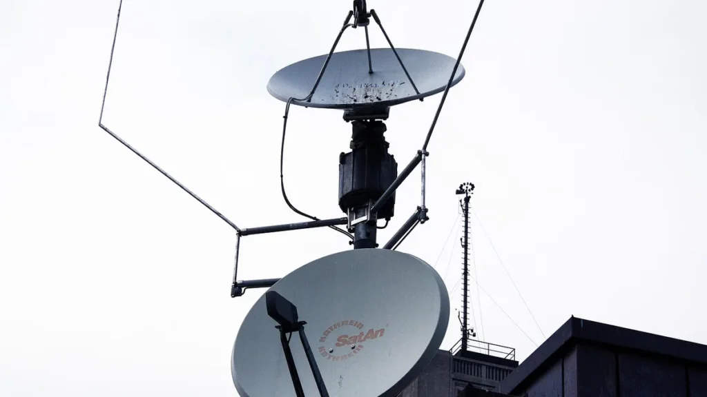 Satellite dish antennas mounted on a structure with a communications tower visible in the background under a cloudy sky.