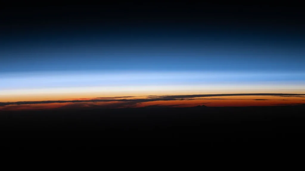 A vibrant sunset horizon showcases colorful orange clouds, blue atmospheric layers, and the dark expanse of space above.