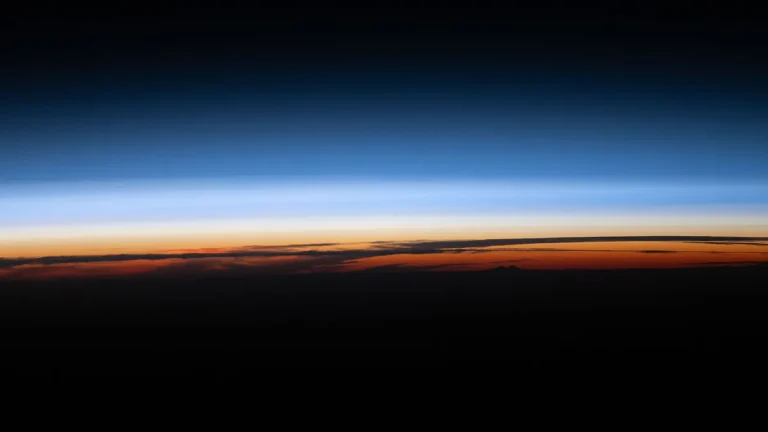 A vibrant sunset horizon showcases colorful orange clouds, blue atmospheric layers, and the dark expanse of space above.