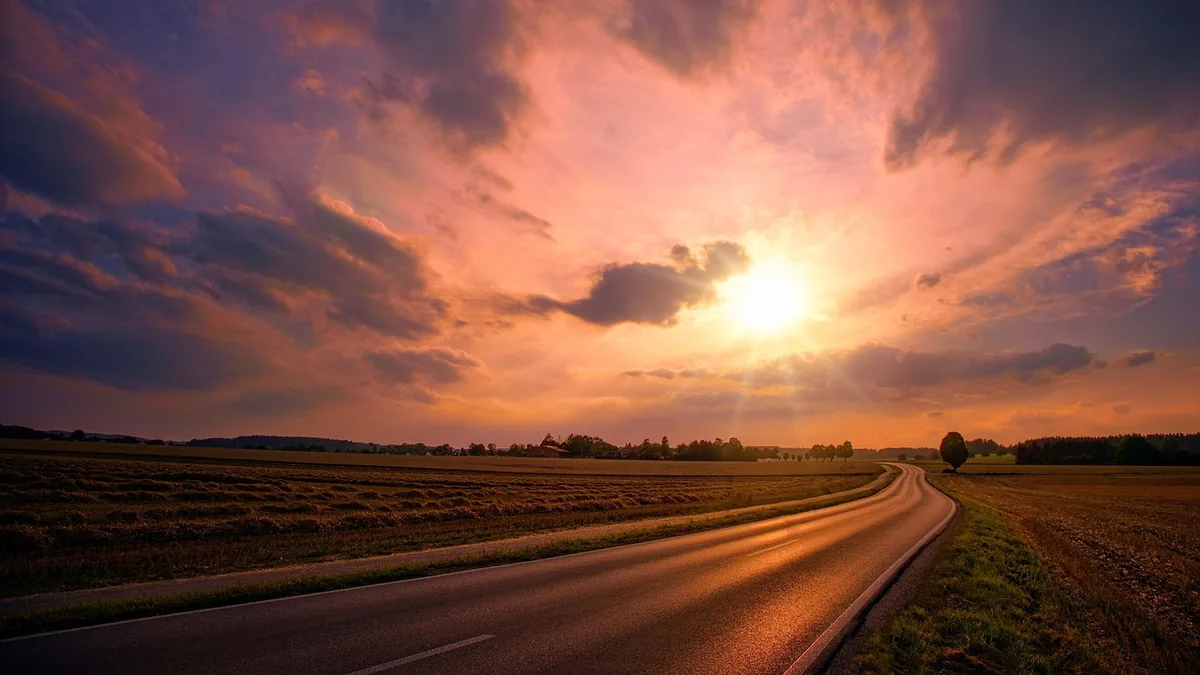Vibrant orange sunset over a winding road and golden fields, with dramatic clouds filling the colorful evening sky.