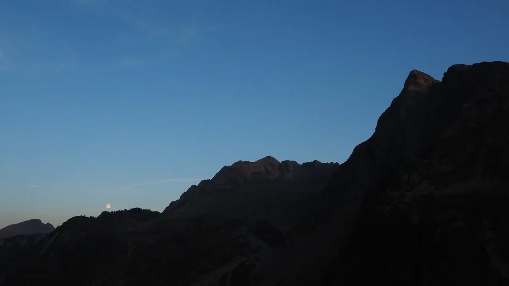 A small moon rises over dark mountain peaks under a clear blue twilight sky.