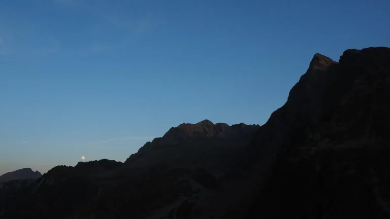A small moon rises over dark mountain peaks under a clear blue twilight sky.