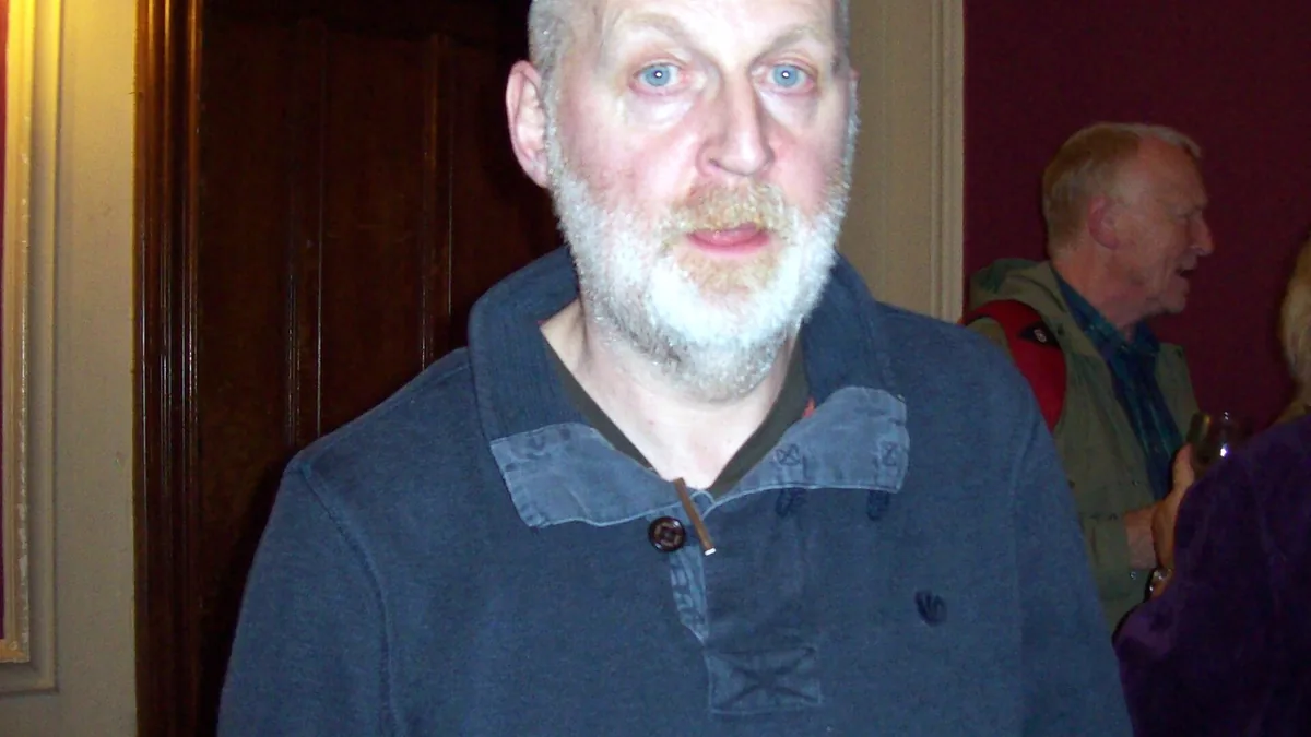 Close-up portrait of a grey-bearded man with blue eyes wearing a dark blue collared sweater.