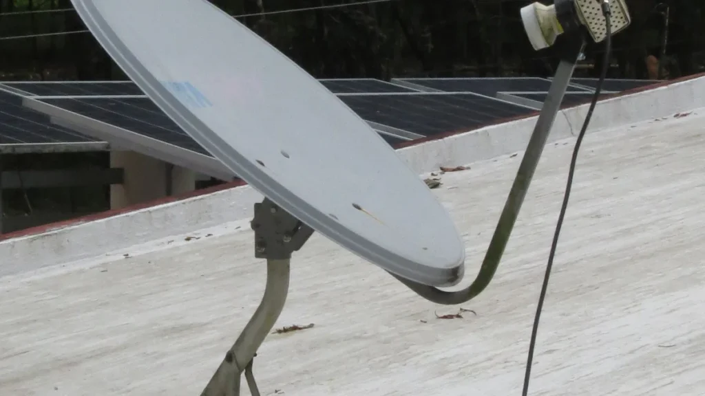 Grey satellite dish on a concrete rooftop with solar panels in the background under soft daylight.