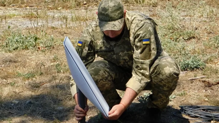 A Ukrainian soldier in camouflage uniform adjusts a Starlink satellite dish in a dry field.