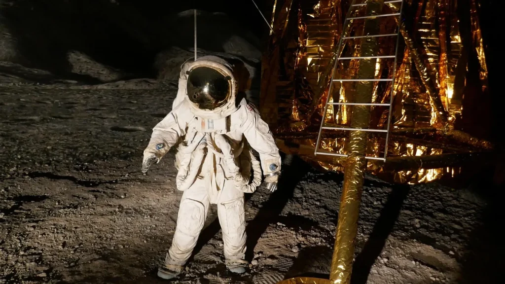 Astronaut in a white spacesuit stands on lunar soil next to a golden lander with a ladder.