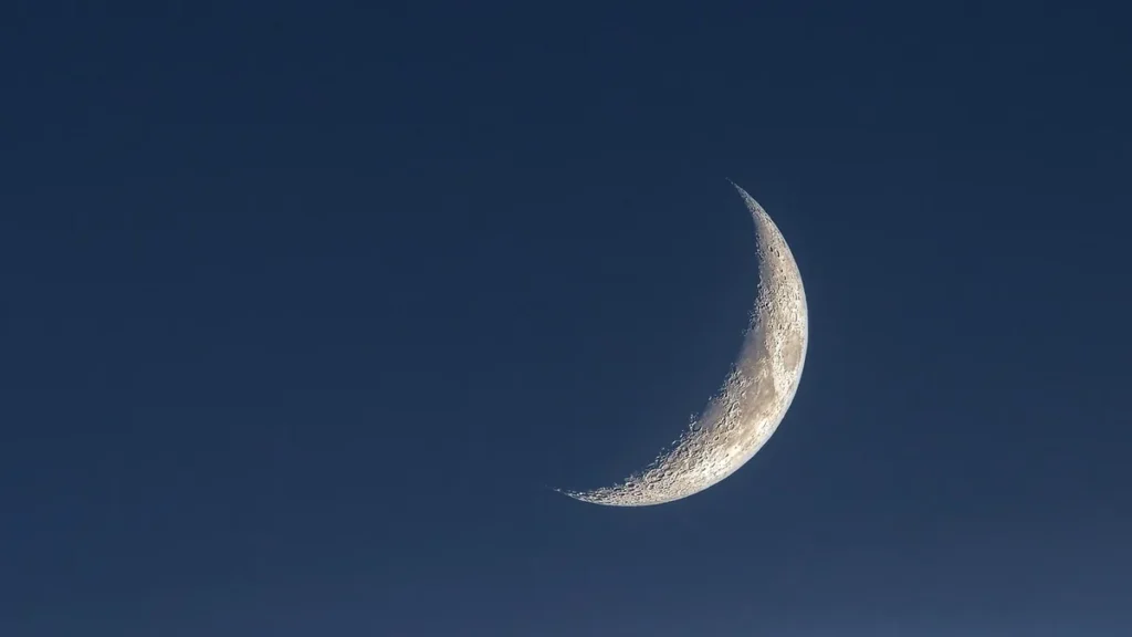 A detailed close-up of a bright crescent moon showing craters against a deep blue sky.