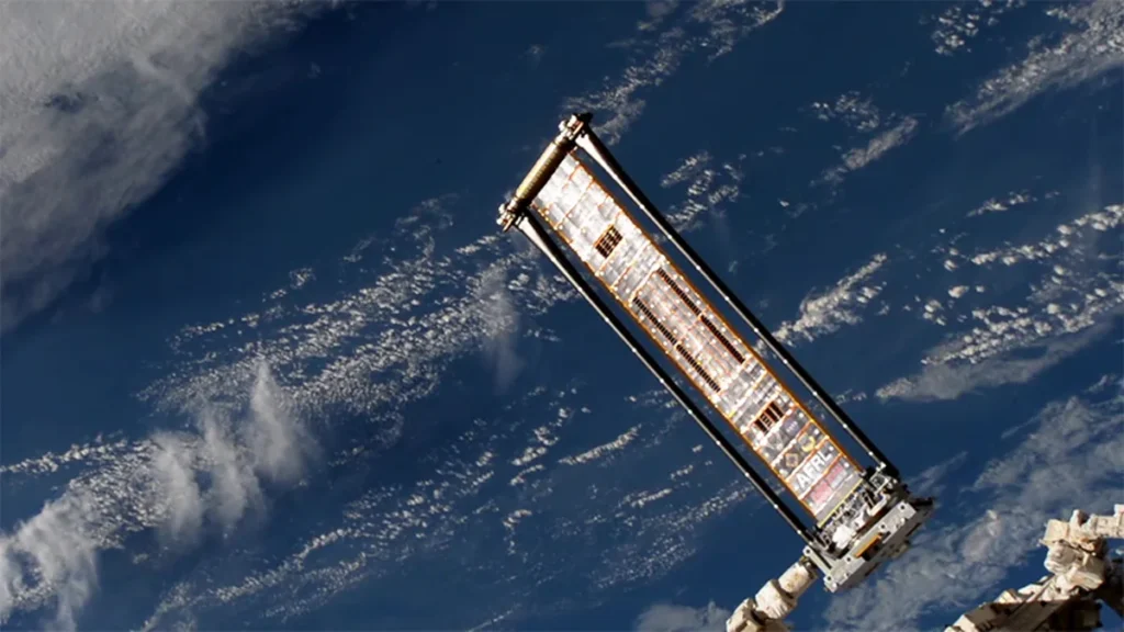 A roll-out solar array panel extended on a robotic arm, with the blue Earth and white clouds below.