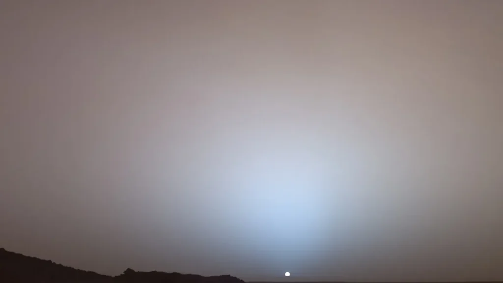 A Martian sunset with a blueish glow around the small sun over a dark horizon.