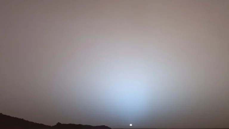 A Martian sunset with a blueish glow around the small sun over a dark horizon.