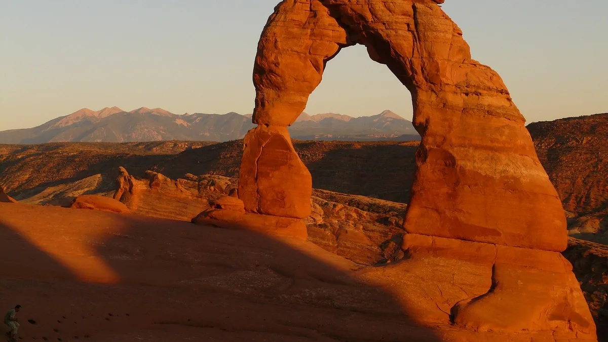 Golden sunset illuminating the iconic red sandstone of Delicate Arch against a mountainous desert backdrop.