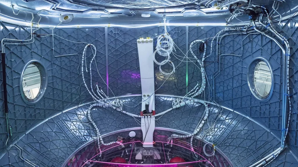 Interior of a futuristic space module with grid walls, circular windows, and colorful wiring.