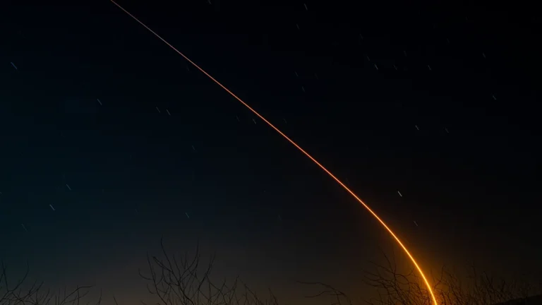 Bright orange space trail curves across a starry night sky toward the horizon behind bare trees.