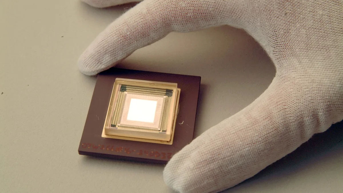 A gloved hand holds a square scientific sensor chip with a bright, reflective central core.