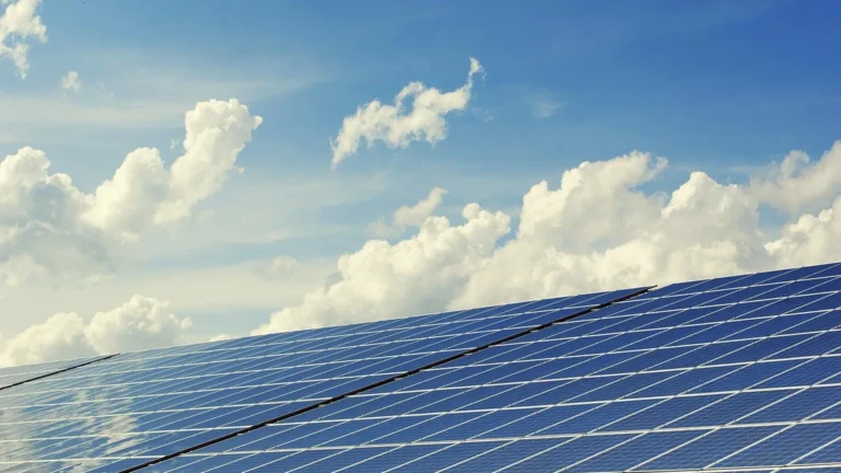 Blue solar panels aligned in rows under a bright blue sky with white clouds.