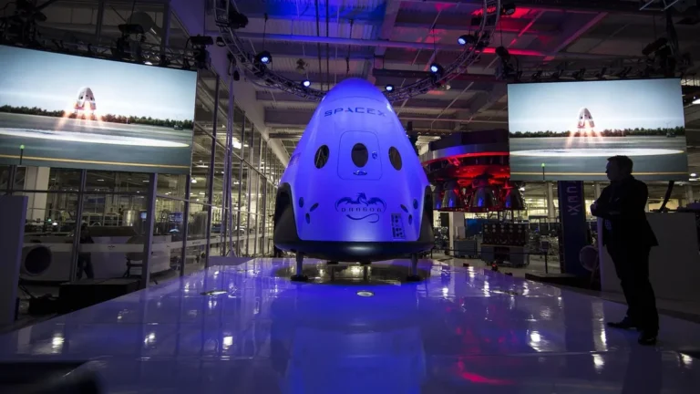 SpaceX Dragon capsule on display under vibrant blue lighting with a man observing the spacecraft.