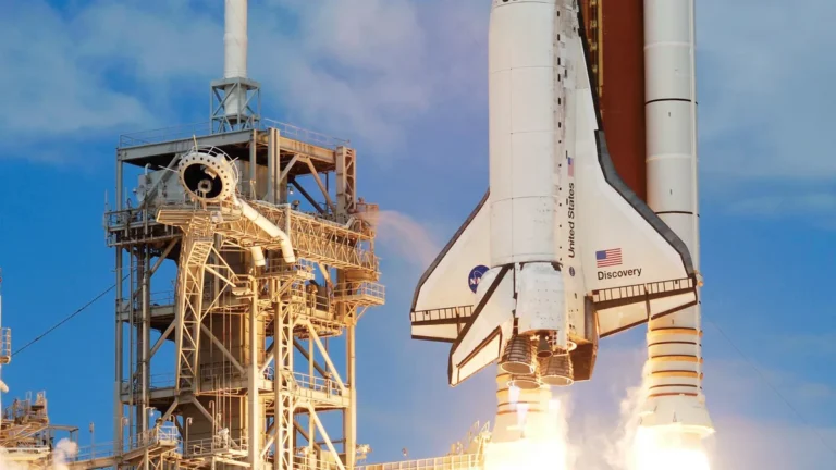 Space Shuttle Discovery launches with bright flames next to the launch tower under a blue sky.
