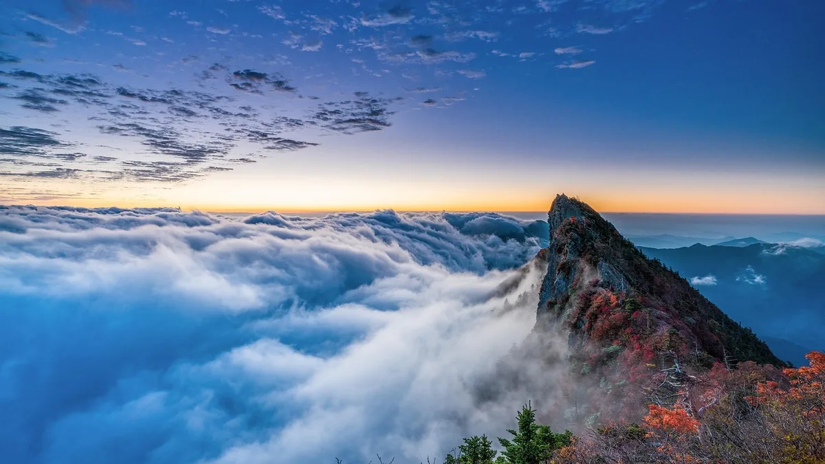 A mountain peak with vibrant autumn foliage rises above a sea of clouds under a colorful twilight sky.