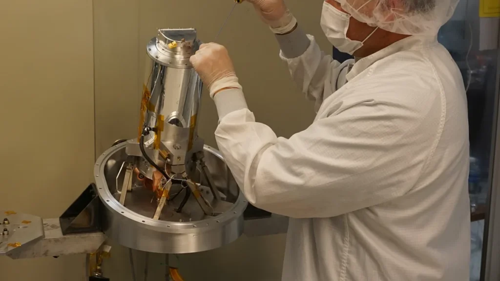 Technician in a cleanroom suit assembling a metallic cylindrical satellite component in a lab setting.