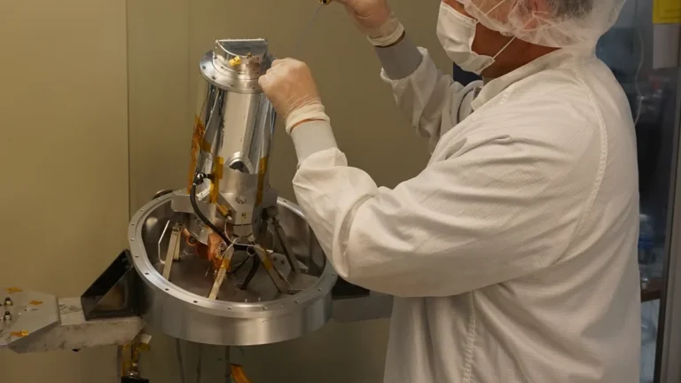 Technician in a cleanroom suit assembling a metallic cylindrical satellite component in a lab setting.
