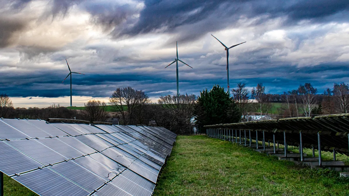 Rows of solar panels in a grassy field with wind turbines standing against a dramatic, cloudy sky.