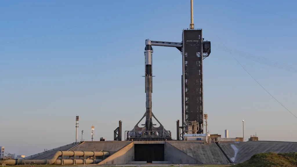 A SpaceX Falcon 9 rocket with a Crew Dragon capsule stands on the launch pad under a clear blue sky.