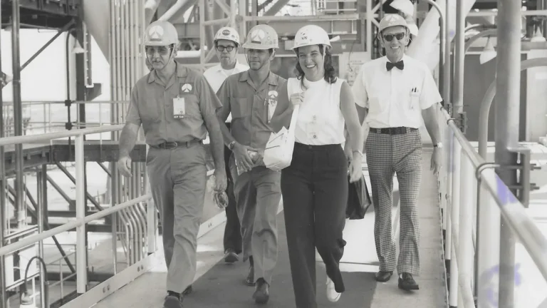 Historical black and white photo of NASA engineers walking on a gantry bridge wearing hard hats.