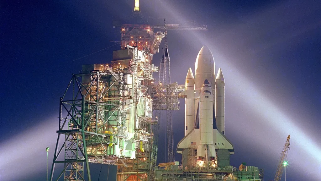 Space shuttle Columbia on launch pad illuminated by bright searchlights at night.