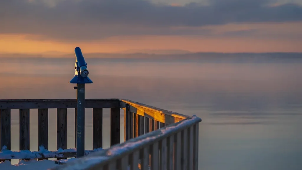 A blue telescope on a snowy wooden pier overlooking a misty lake at sunset.
