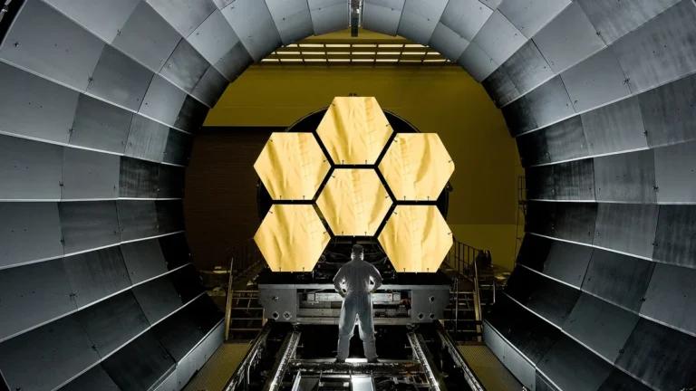 Person in flight suit inspecting a large hexagonal telescope structure inside a large chamber, likely part of a space or