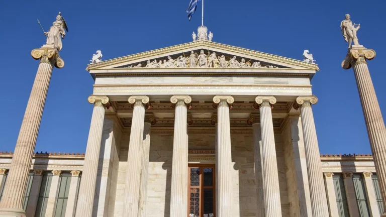 The grand neoclassical facade of the Academy of Athens with columns and statues under a blue sky.