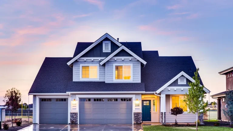 A modern grey two-story house with a double garage lit up under a colorful twilight sky.