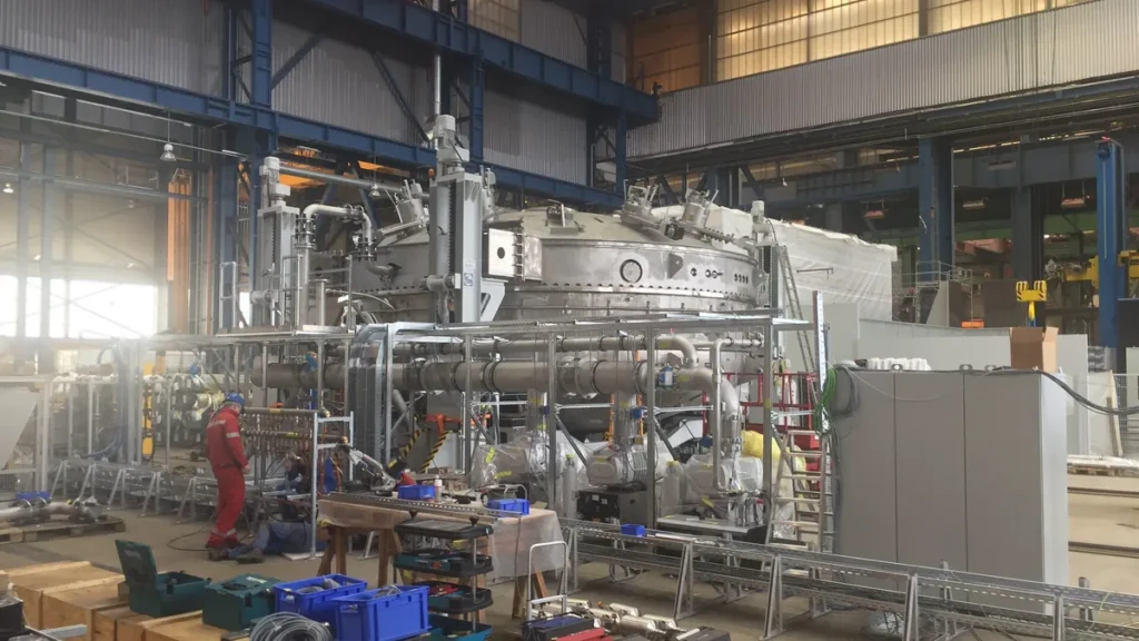 Large silver scientific machinery with pipes and scaffolding inside an industrial workshop.