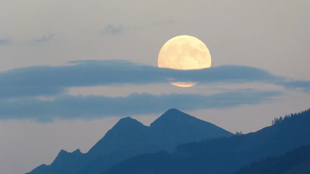 Large golden full moon partially obscured by clouds, rising over silhouetted mountain peaks in twilight sky.