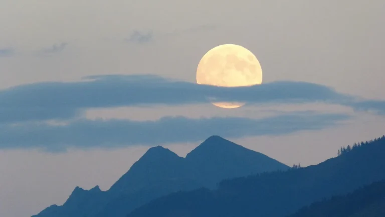 Large golden full moon partially obscured by clouds, rising over silhouetted mountain peaks in twilight sky.