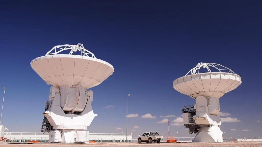 Two giant white radio telescope antennas under a clear blue sky, with a white pickup truck parked between them.