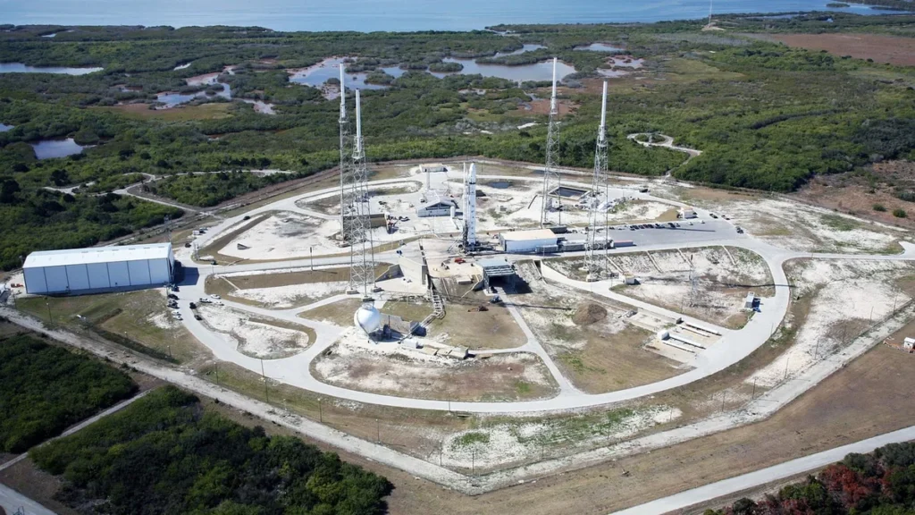 A white rocket stands on a launch pad surrounded by four tall lightning towers, set against a green coastal landscape.