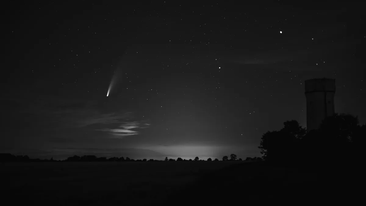 Bright comet with a glowing tail shines in a starry black and white night sky over a silhouetted landscape.