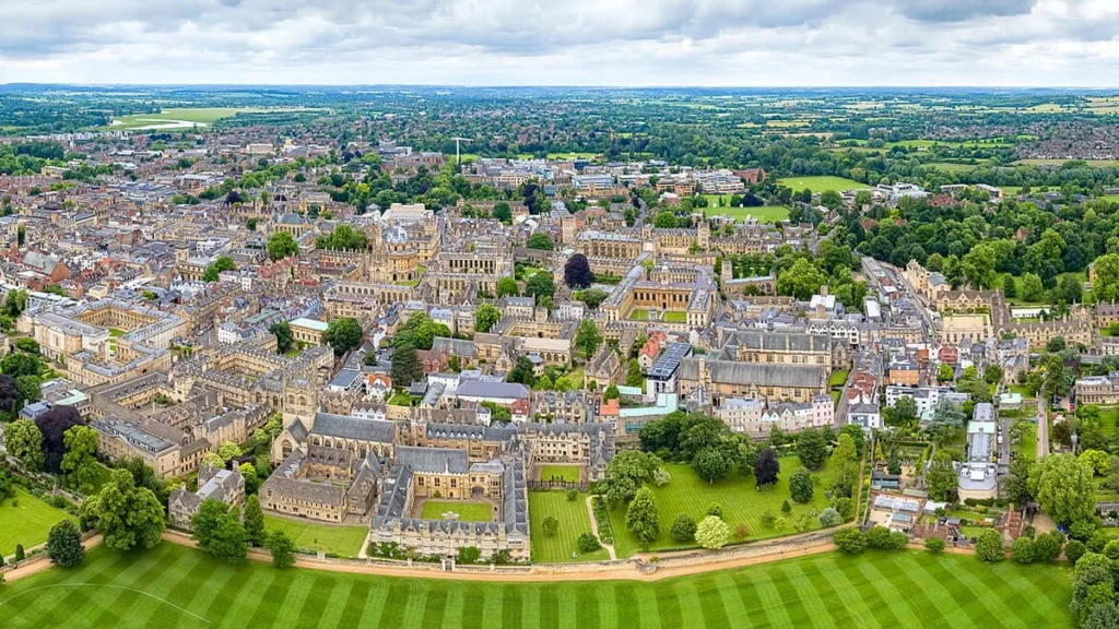 Aerial view of historic Oxford city and colleges under a cloudy sky, with green lawns in the foreground.