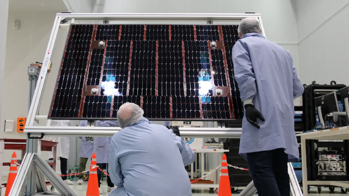 Technicians in protective clothing inspect a large solar panel array mounted on a test rig in a cleanroom.