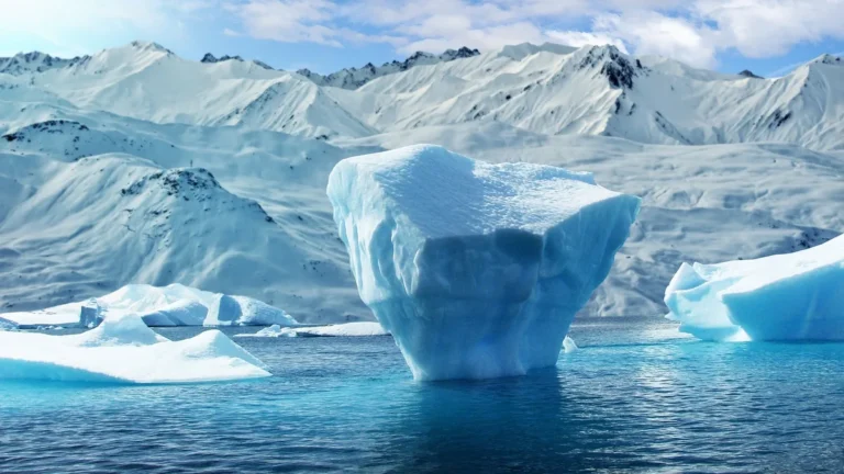 A majestic iceberg floats in blue water against a backdrop of snow-covered mountains under a partly cloudy sky.